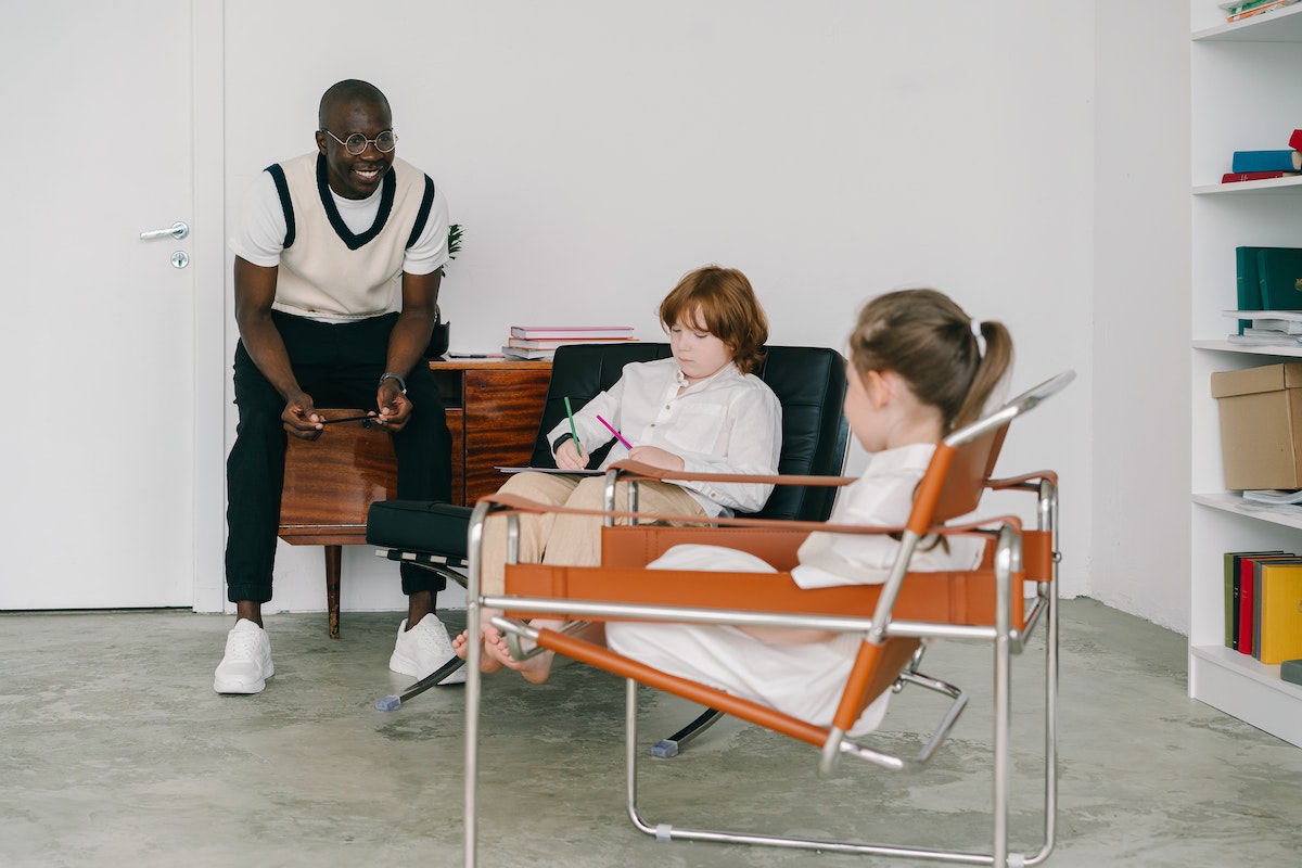 Um profissional falando com duas crianças autistas que estão sentadas, cada uma em uma cadeira