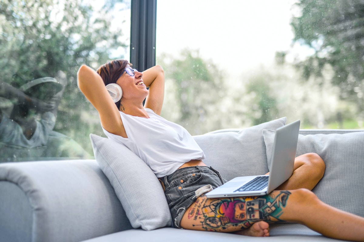 Uma mulher sentada em seu sofá. Ela está com o notebook em seu colo, um fone de ouvido e tem tatuagem na perna
