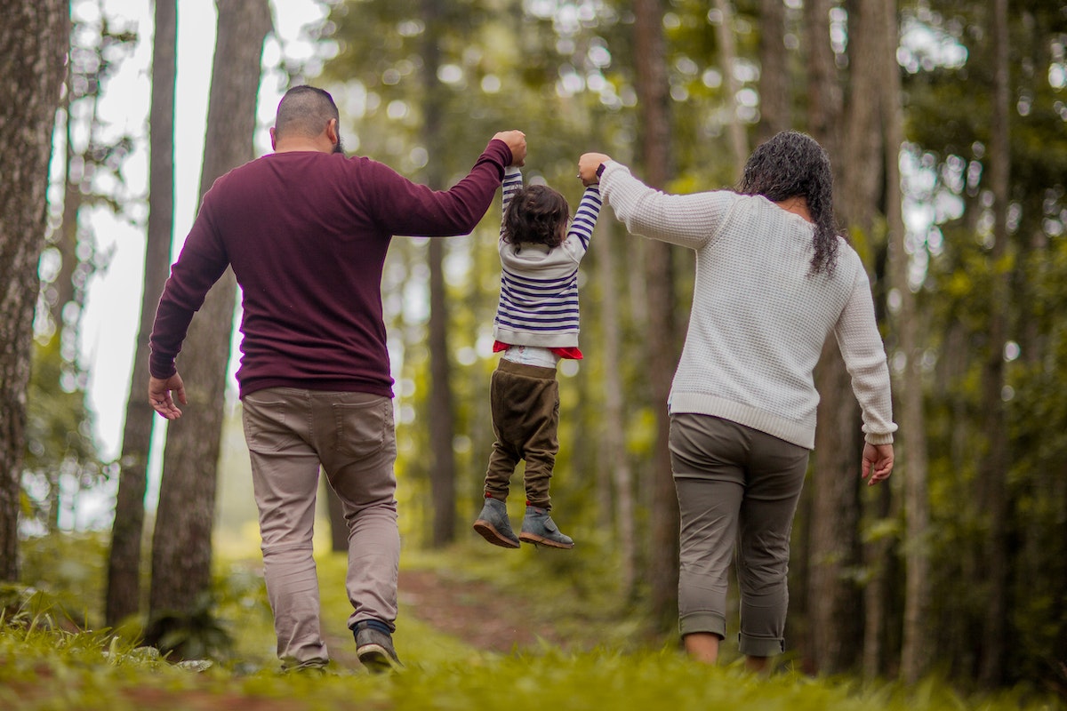 Foto de pais segurando a criança pelos braços. Eles estão em um bosque com muitas árvores