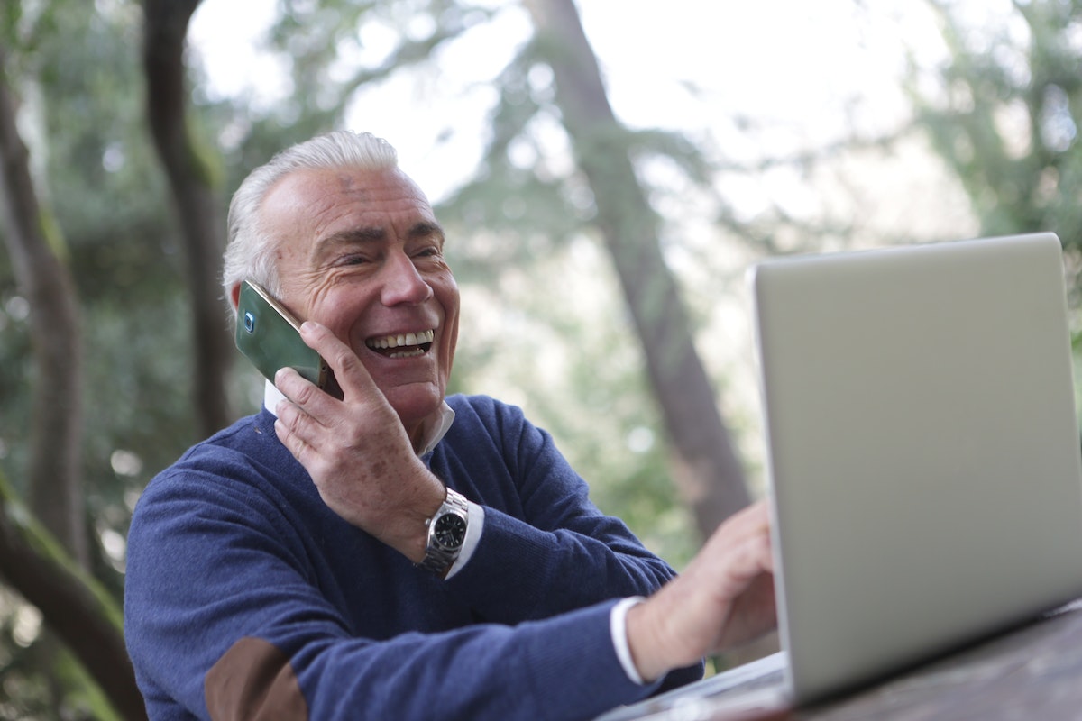 Um senhor muito feliz confere o Piso INSS 2024 no notebook enquanto fala em seu celular