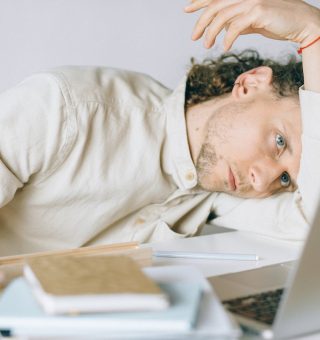 Homem com a cabeça encostada no braço. Ele está com cara de cansado e olha para o notebook enquanto confere os Direitos INSS para Pessoas com Síndrome de Burnout 2024